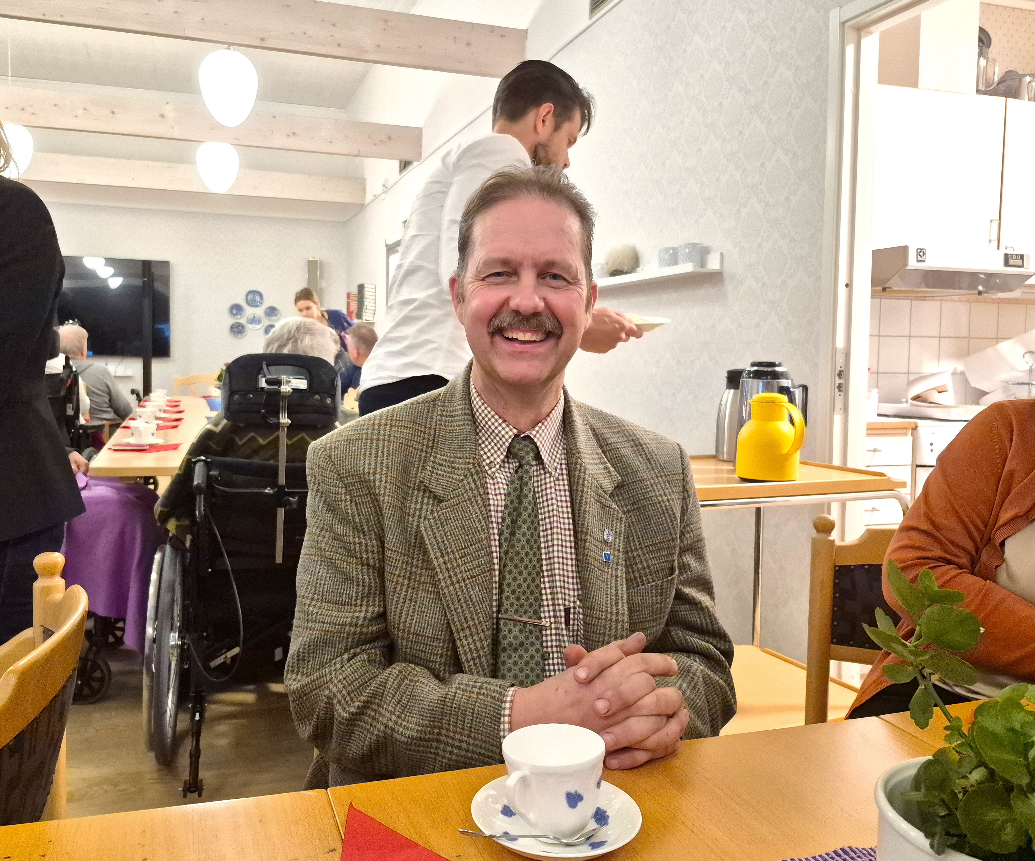 Daniel Jensen sitter och ler med en kopp kaffe på bordet framför sig. Han sitter tillsammans med de boende och fikar och har det trevligt.