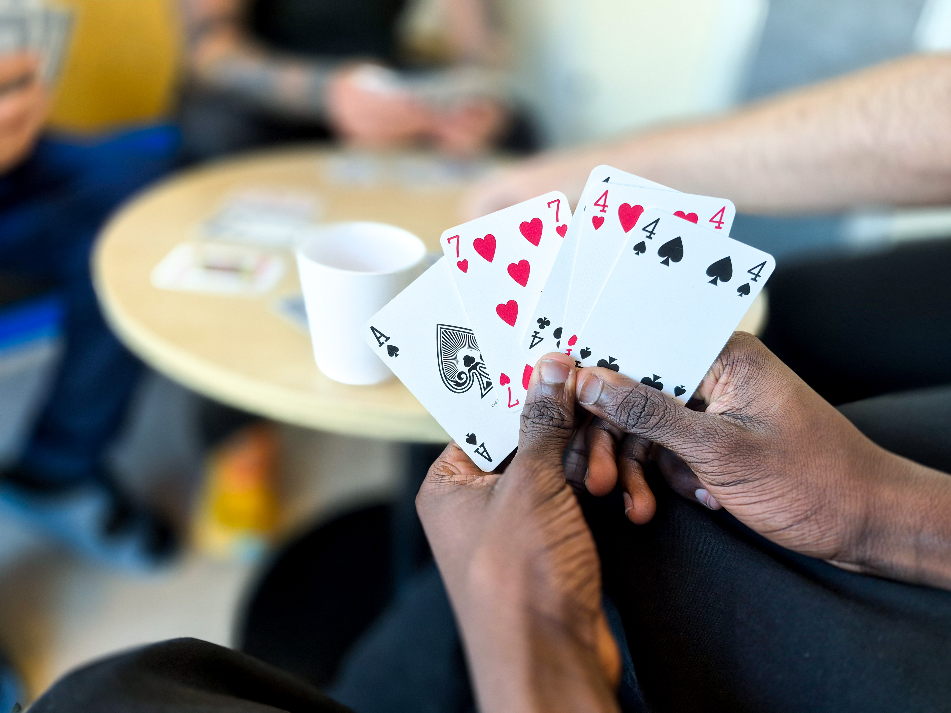 Händer som håller i några spelkort. I bakgrunden syns ett bord med fler personer runt och fler spelkort, men detta är ur fokus.