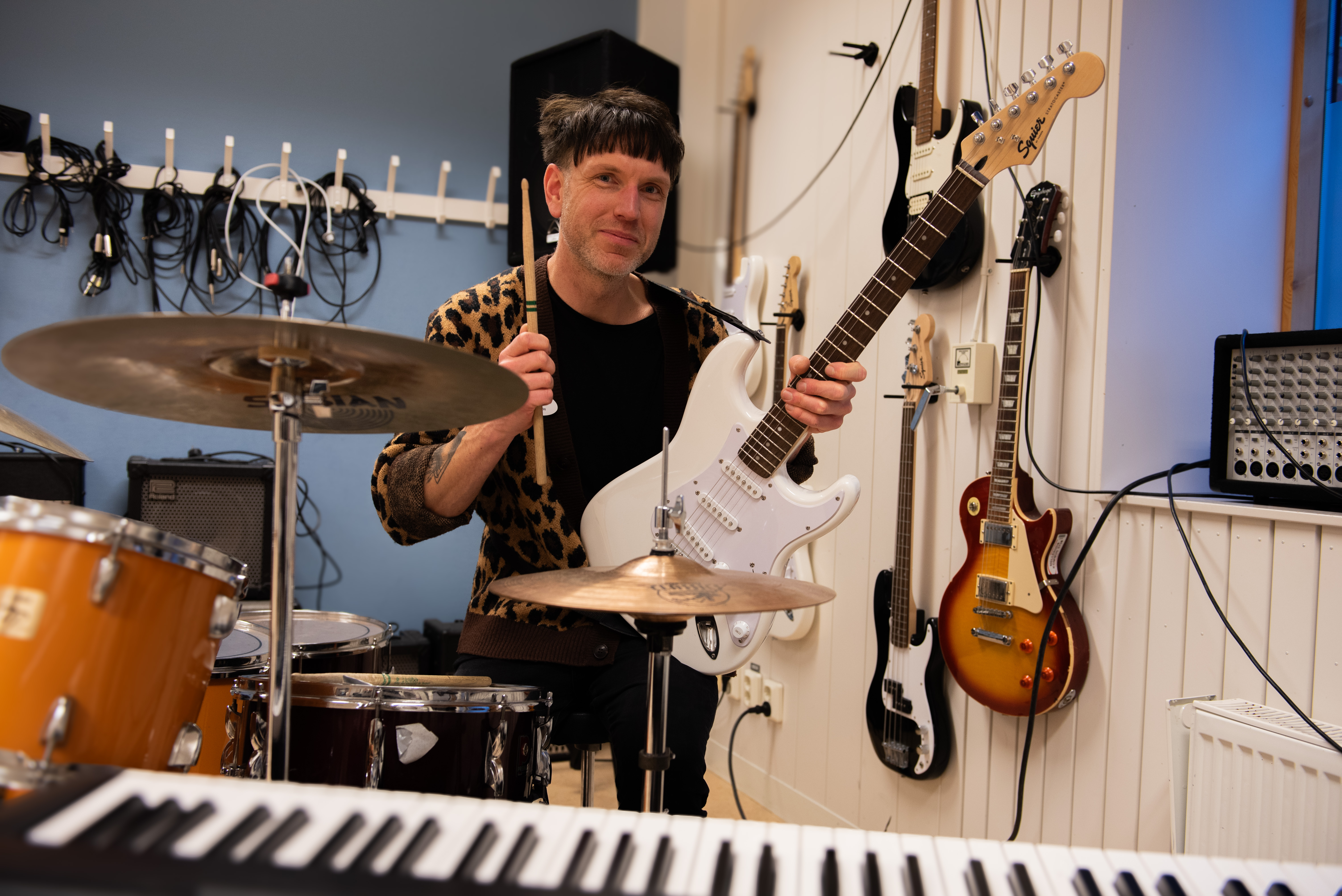 Foto på en glad musiklärare vid ett trumset, hållandes i en gitarr och trumpinne.