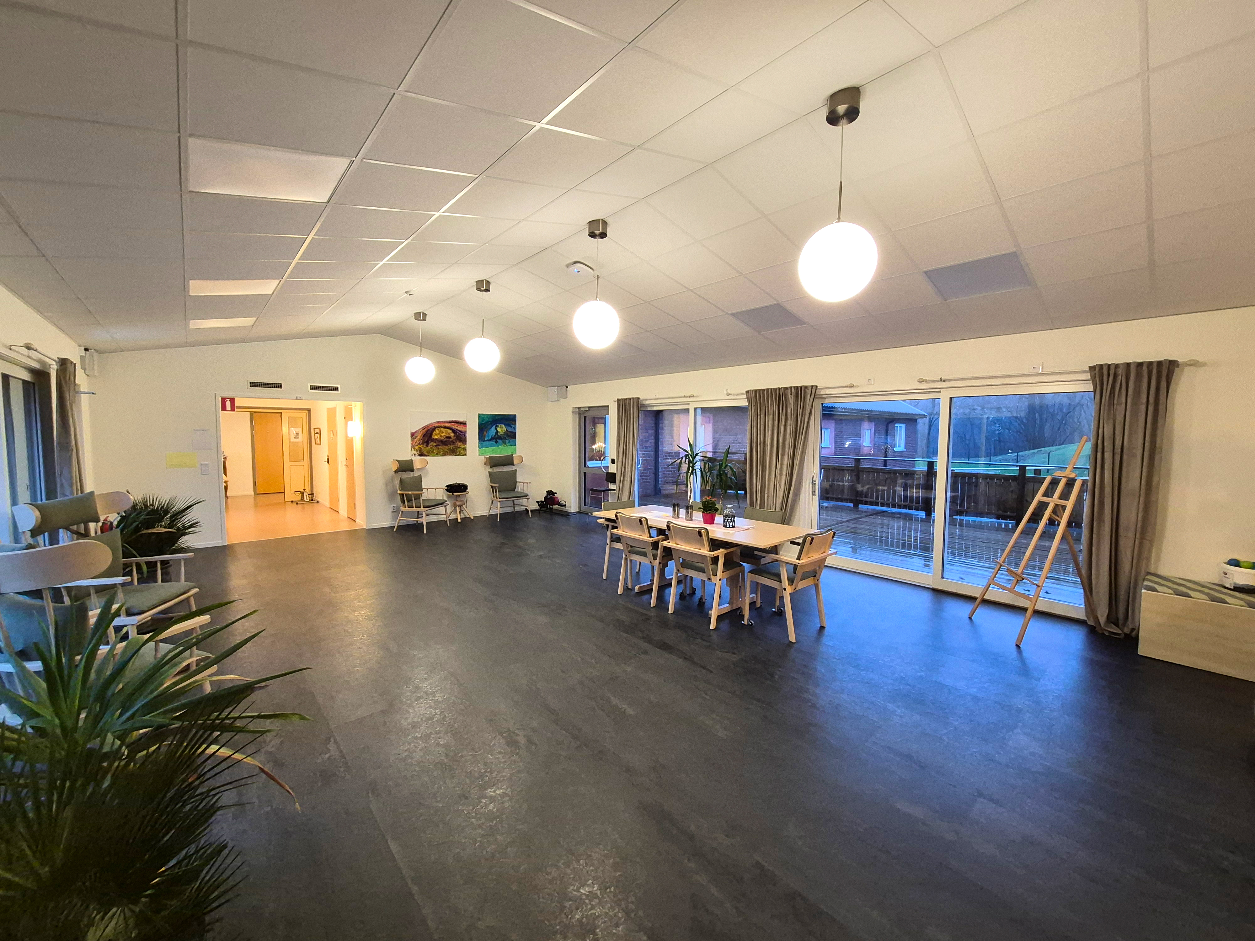 Vinterträdgård med stengolv, stora glaspartier på väggarna som kan öppnas upp och flera gröna växter i det stora och ljusa rummet.