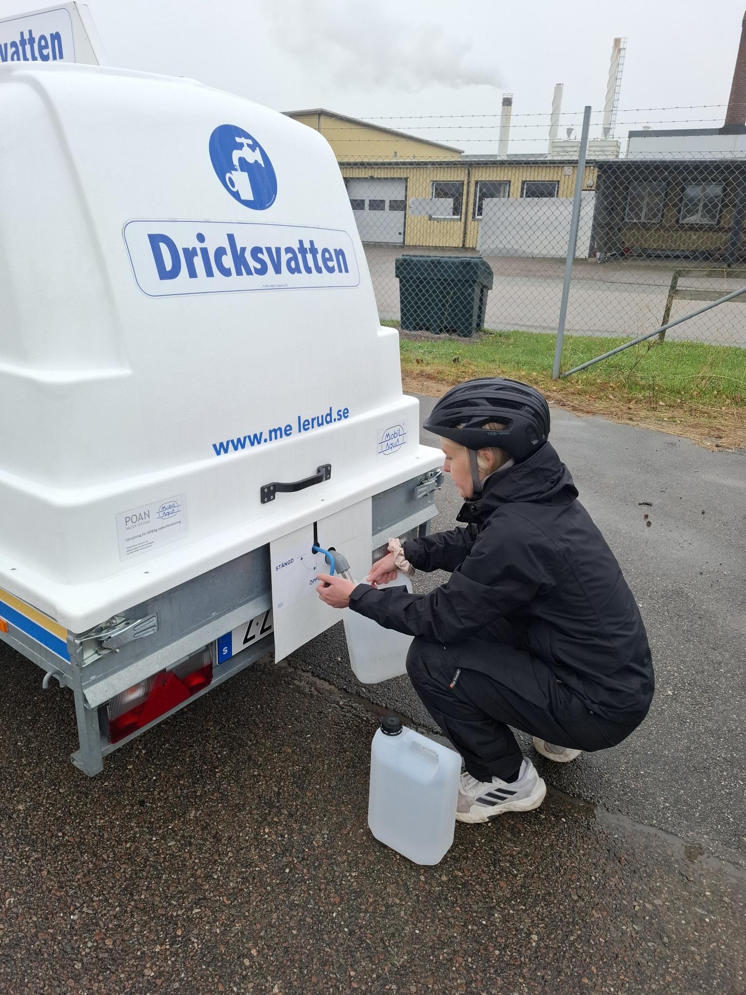 Person hukar sig bakom ett släp som det finns dricksvatten på och fyller på en dunk från en kran på släpet.