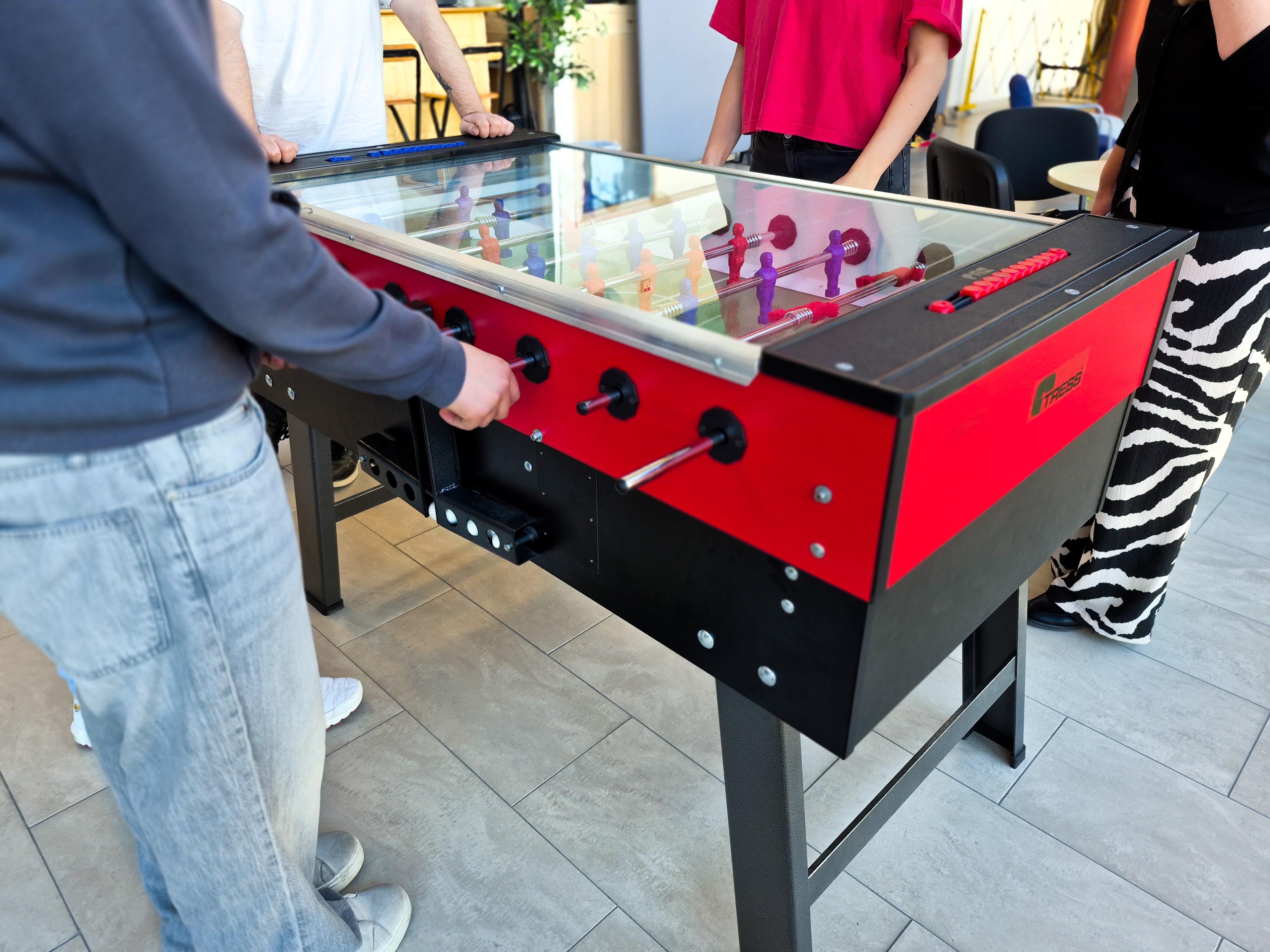 4 personer står runt ett analogt fotbollsspel där det är flera figurer på pinnar som man snurrar på för att försöka få i bollen i motståndarens mål